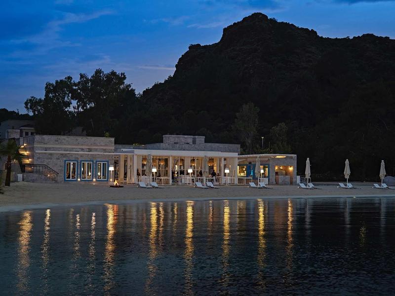 Restaurace u vody osvětlená večer s horou v pozadí.