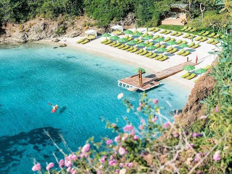 Pláž s bílým pískem, lehátky a tyrkysovou vodou v zátoce obklopené zelení.