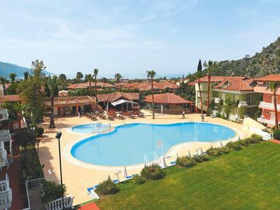 Hotel complex with large pool, sun umbrellas, and red roofs in a mountainous landscape.
