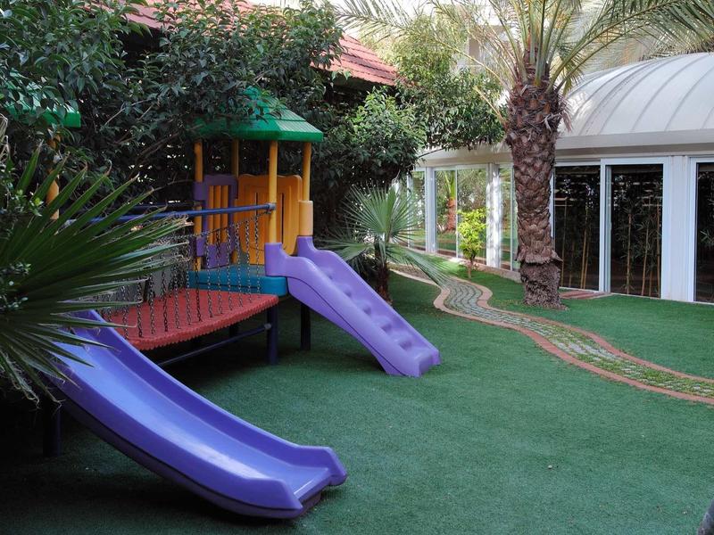 Area giochi con due scivoli viola e prato verde artificiale davanti a un edificio con finestre in vetro.