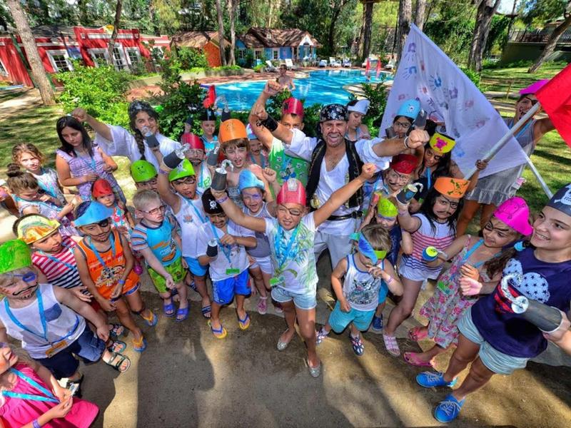 Gruppenkinder und Betreuer in bunter Kleidung feiern draußen auf einem Spielplatz bei Sonnenschein.