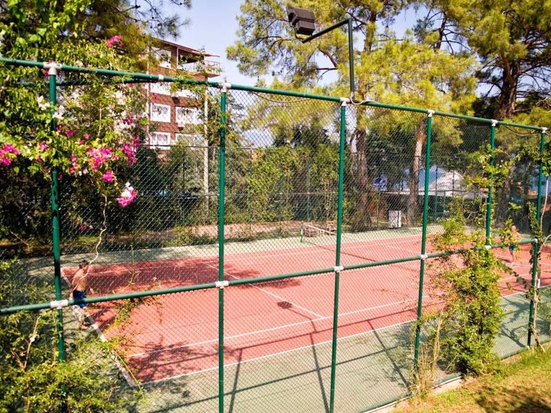 Roter Tennisplatz umgeben von grünem Metallzaun und Bäumen bei sonnigem Wetter.