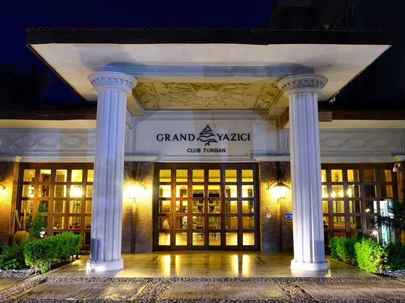 Eingang eines Gebäudes mit zwei weißen, korinthischen Säulen und beleuchteten Holztüren bei Nacht.