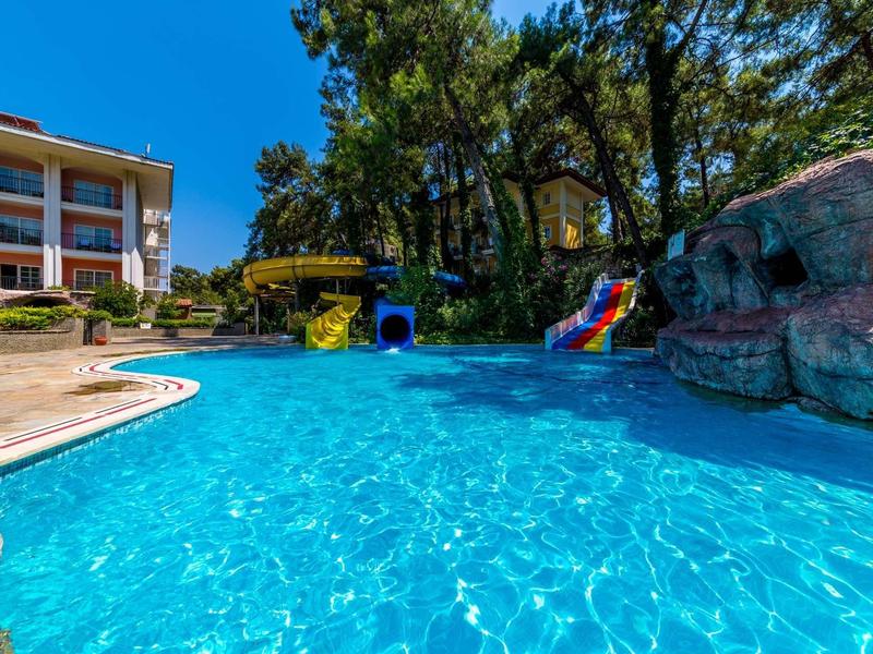 Großes Schwimmbecken mit klarem Wasser, Rutschen, Felsen und einem mehrstöckigen Gebäude im Hintergrund.