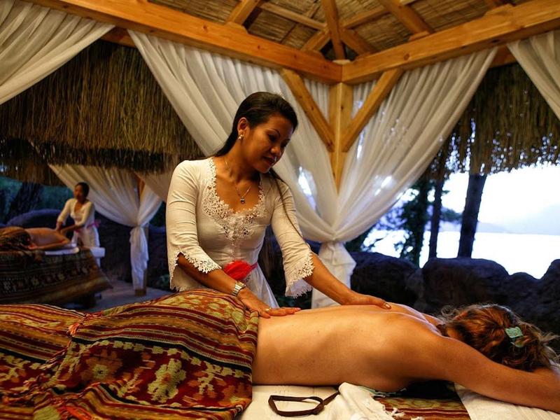 Frau massiert einen Mann auf einem bunten Tuch in einem offenen, hölzernen Pavillon am Wasser.
