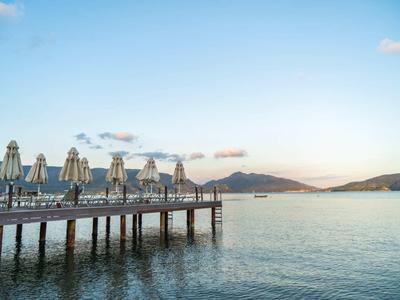 Pier with closed umbrellas over calm sea under clear sky with hills in the background.
