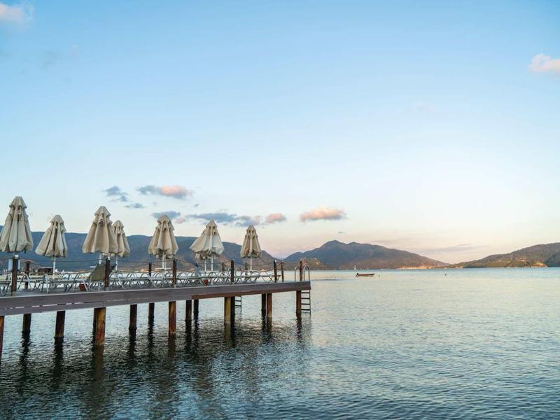 Pier with closed umbrellas over calm sea under clear sky with hills in the background.