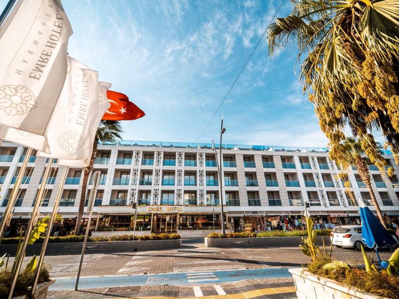 Modern hotel building with many windows, flags, and palm trees under a blue sky.