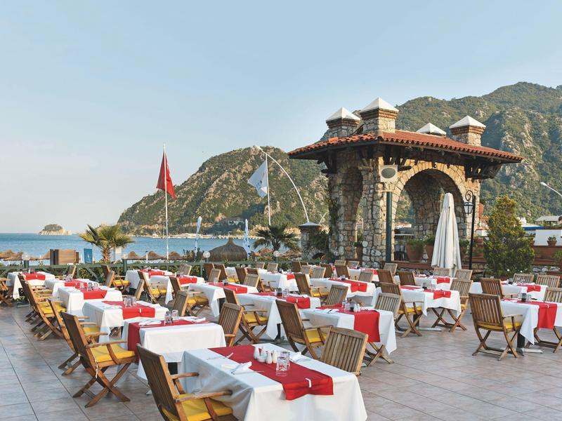 Restaurant en plein air avec des tables dressées offrant une vue sur la mer et les collines en arrière-plan.