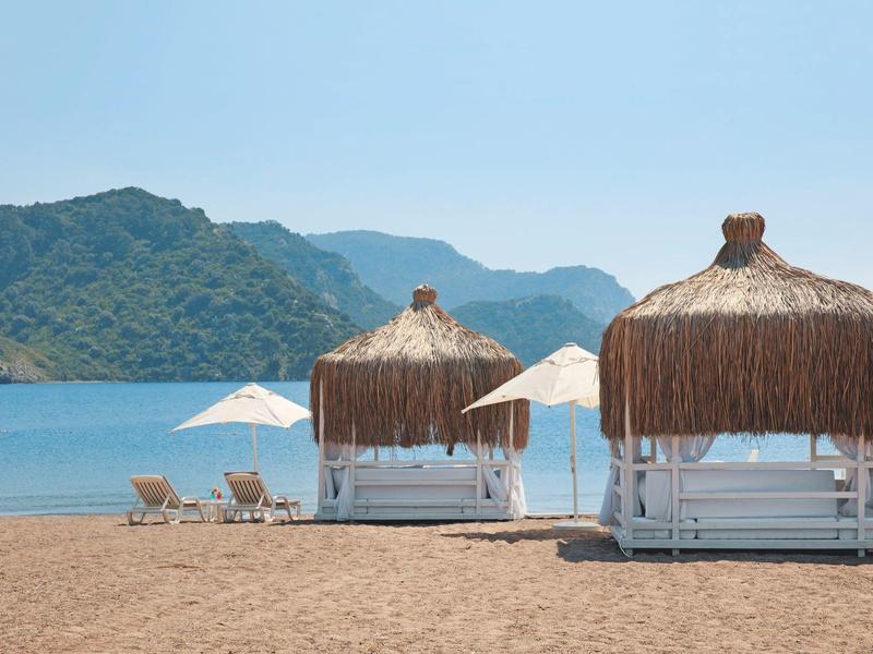 Plage avec deux cabanons en paille, parasols blancs et chaises longues avec montagnes et ciel clair.