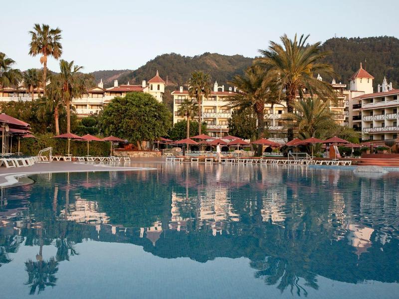 Grand bassin de l'hôtel avec chaises longues et palmiers devant un bâtiment à plusieurs étages et des montagnes.
