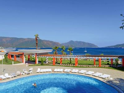 Runder Pool mit Liegestühlen, dahinter Blick auf blaue Berge und das Meer bei klarem Himmel.