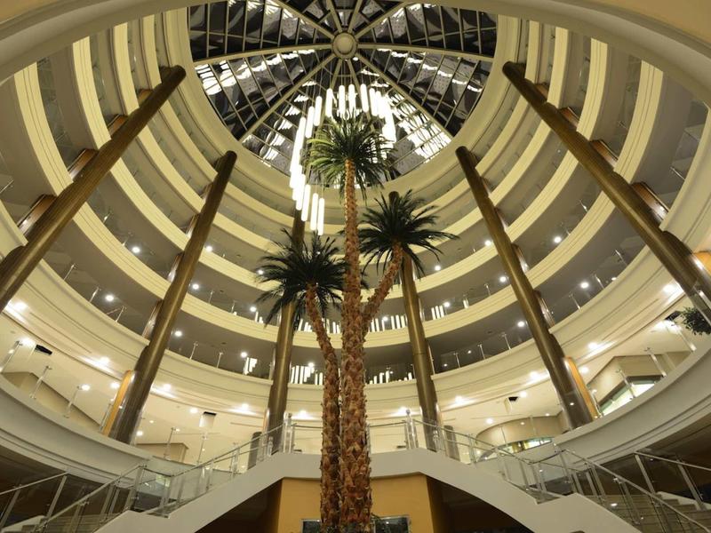 Runde Hotellobby mit hohen Palmen, Glasdach und mehrstöckigen Balkonen im Innenbereich.
