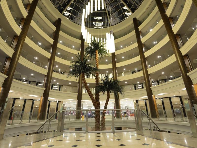 Innenansicht eines runden Atriums mit Glasdach, Palmen und mehrstöckigen Balkonen.