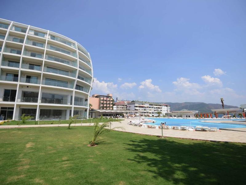 Modernes rundes Hotelgebäude mit Pool, Liegestühlen und Grünfläche unter blauem Himmel.