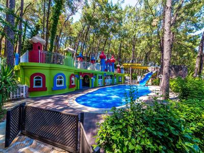 Parque acuático colorido con piscina y tobogán, rodeado de árboles y vegetación verde.