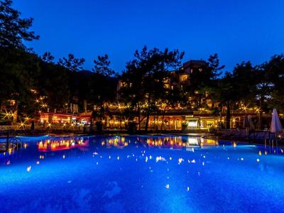 Piscina iluminada frente a un hotel por la noche con árboles y cielo azul.