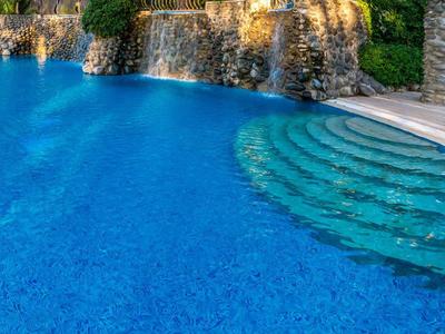 Einladender blauer Pool mit Rundtreppen und Wasserfall vor grünem Hintergrund.