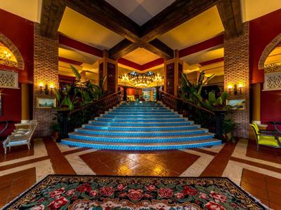 Elegante recepción con escalera iluminada, plantas y alfombra roja en un hotel.