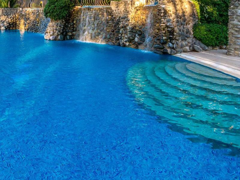 Piscina azul de hotel con escalones redondos y cascada rodeada de naturaleza y columnas.