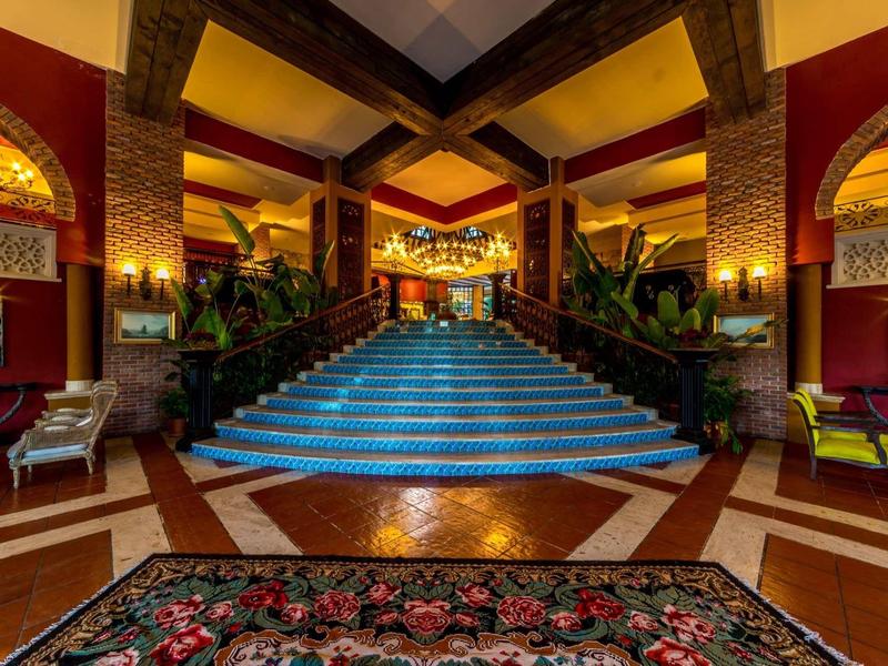 Elegante recepción con escalera iluminada, plantas y alfombra roja en un hotel.