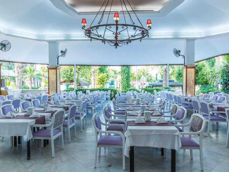 Sala da pranzo luminosa con tavoli apparecchiati e sedie viola, circondata da finestre di vetro.