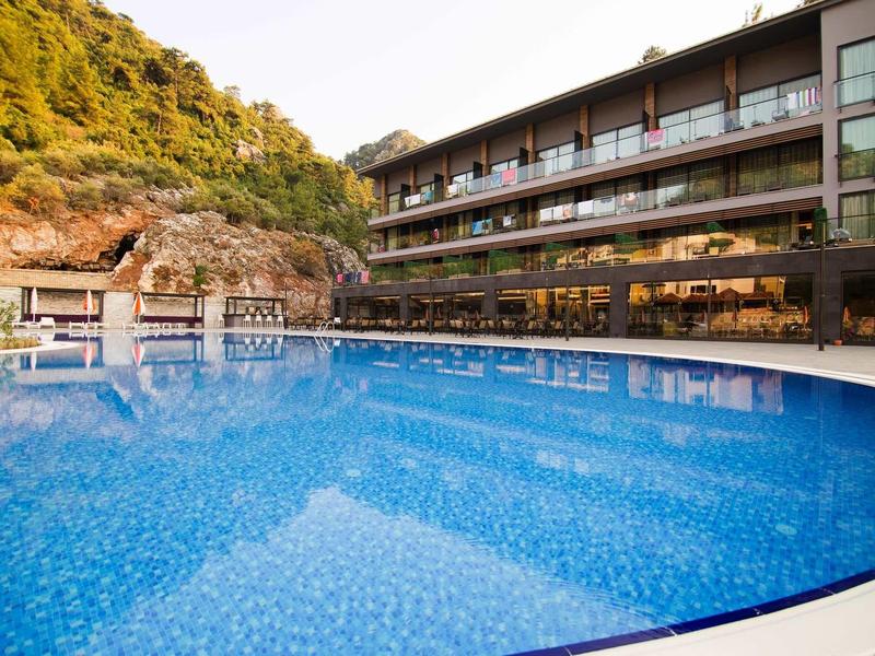 Großer runder Pool mit blauem Wasser vor einem modernen Hotelgebäude und bewaldetem Hang im Hintergrund.