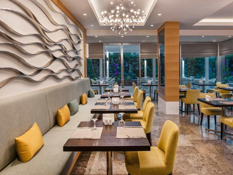 Modernes Restaurant mit gelben Stühlen, Holz- und Glas-Tischen, dekorativer Wand und großen Fenstern.