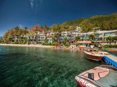 Heldere waterkant met hotel en boten onder een blauwe lucht en beboste heuvels.