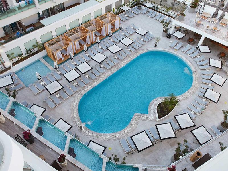 Modern hotel pool area with sun loungers, cabanas, and nearby palm trees in a sunny setting.