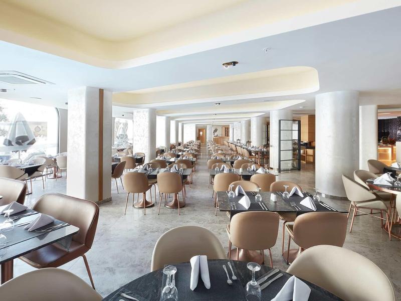 Spacious hotel dining room with many tables and beige chairs under soft lighting.