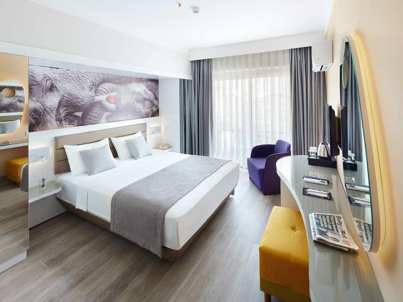Modern hotel room with double bed, wall art, desk, chair, and large window with curtains.