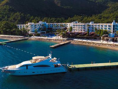 Yacht de luxe amarré près d'un hôtel sur une plage côtière avec des montagnes verdoyantes en arrière-plan.