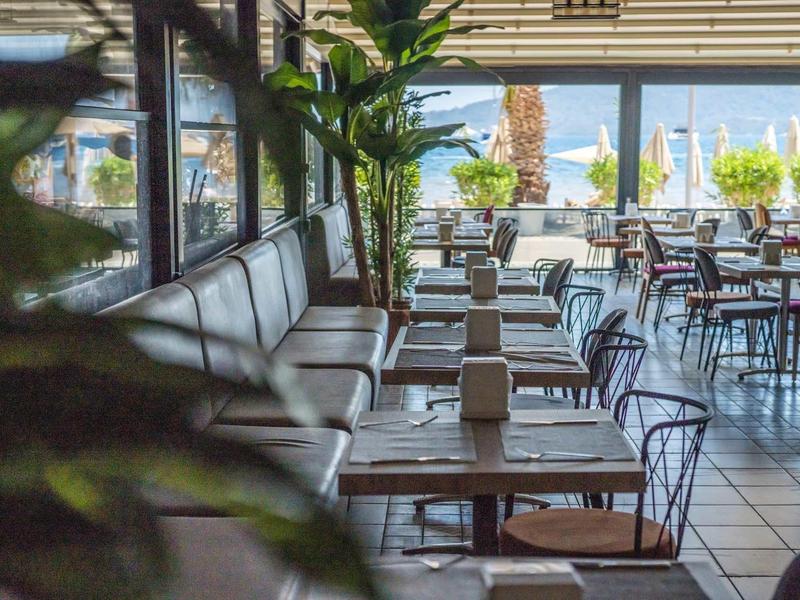 Moderní hotelová restaurace s lavicemi a židlemi, velkými okny a výhledem na palmy.