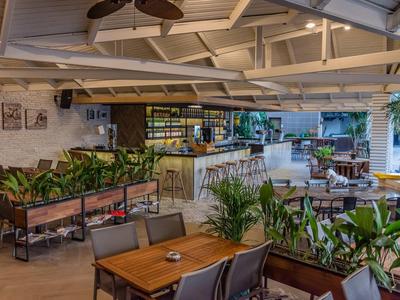 Licht hotelrestaurant met planten, houten tafels en een moderne bar onder een open plafond.