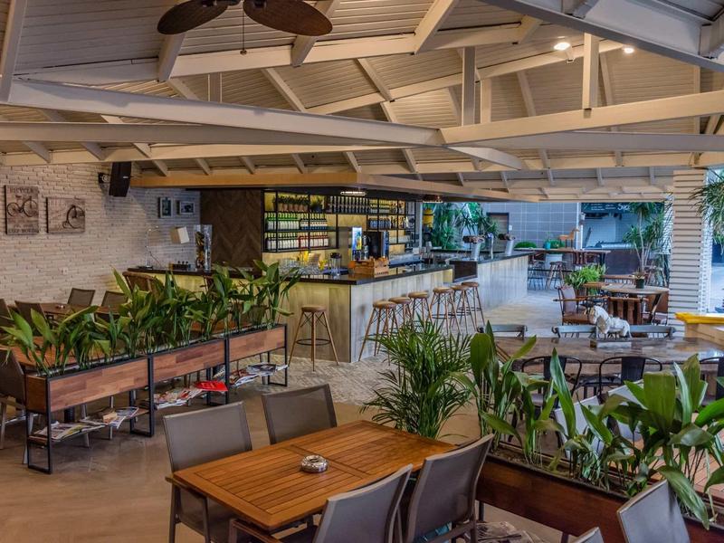 Licht hotelrestaurant met planten, houten tafels en een moderne bar onder een open plafond.