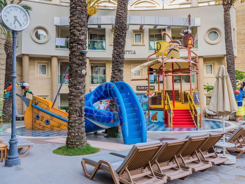 Speelplaats voor kinderen met glijbanen en zitplaatsen naast palmbomen in hotelomgeving.