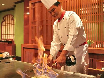 A chef in white uniform prepares a flaming dish on a grill in a restaurant.