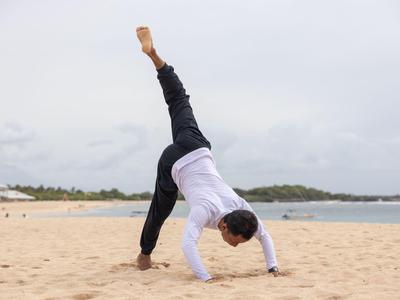 Person macht Yoga mit einem Bein in der Luft am Strand mit Meer im Hintergrund