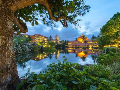 Abendliche Szene mit beleuchtetem Hotel am ruhigen See, umgeben von Bäumen und Pflanzen.