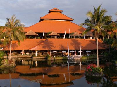 Large three-story hotel building with red roofs beside water and palm trees.