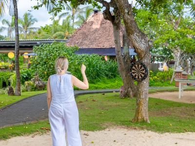 Frau spielt Darts im Garten eines tropischen Resorts mit Palmen und hölzernen Gebäuden im Hintergrund.