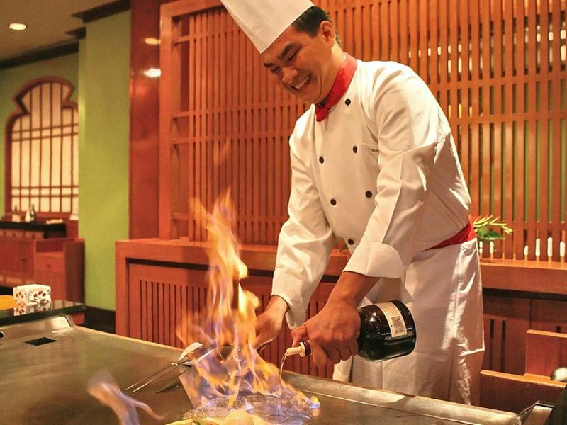 A chef in white uniform prepares a flaming dish on a grill in a restaurant.