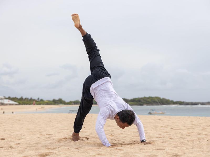 Person practicing yoga with one leg raised on sandy beach under cloudy sky.