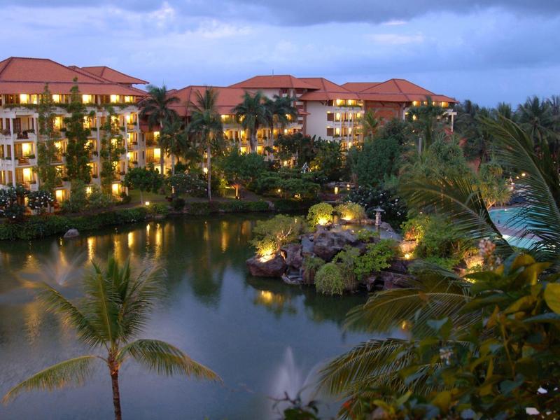 Hotel complex by water with lit buildings and palm trees at dusk.