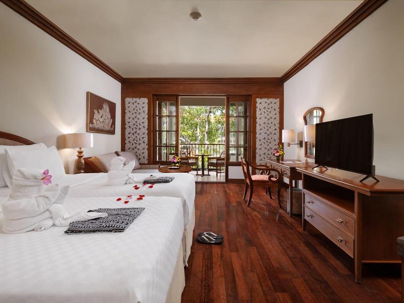 Brightly furnished hotel room with two beds and balcony overlooking garden.