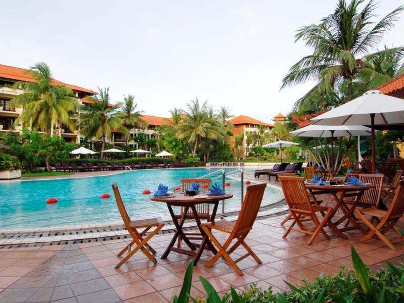 Hotel pool with lounge chairs and umbrellas, surrounded by palm trees and hotel buildings.