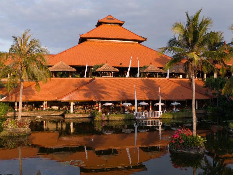 Large three-story hotel building with red roofs beside water and palm trees.