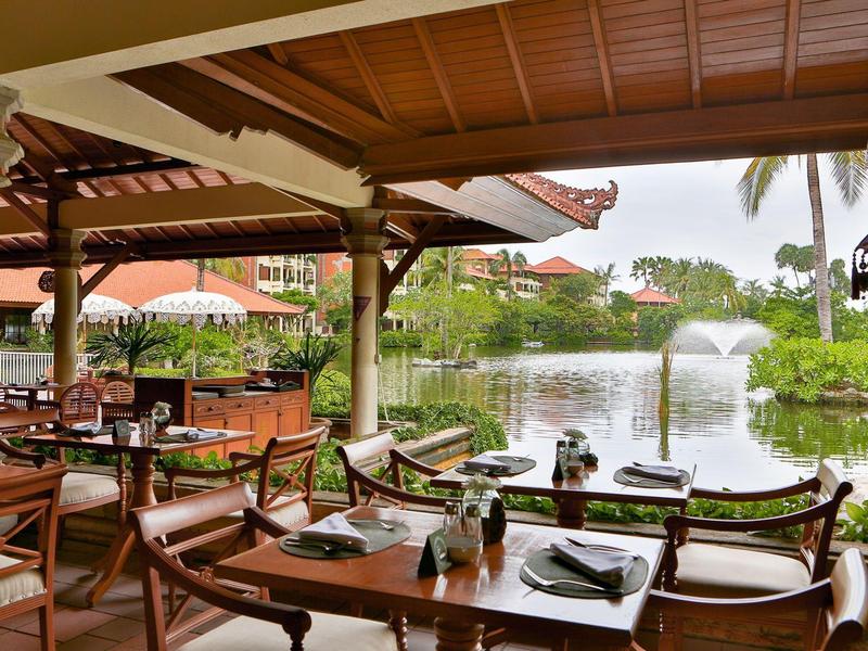 Open restaurant with wooden furniture overlooking a pond with a fountain and tropical vegetation.