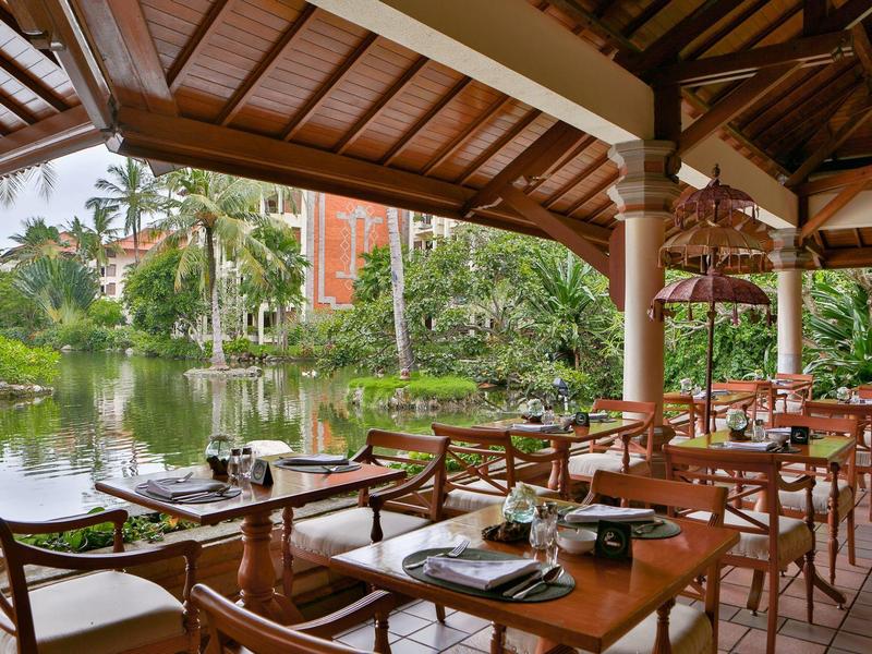 Covered restaurant terrace with wooden tables overlooking a pond with palm trees.